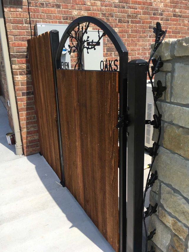 Wooden gate with black metal frame and decorative tree design. Brick and stone walls surround the gate.