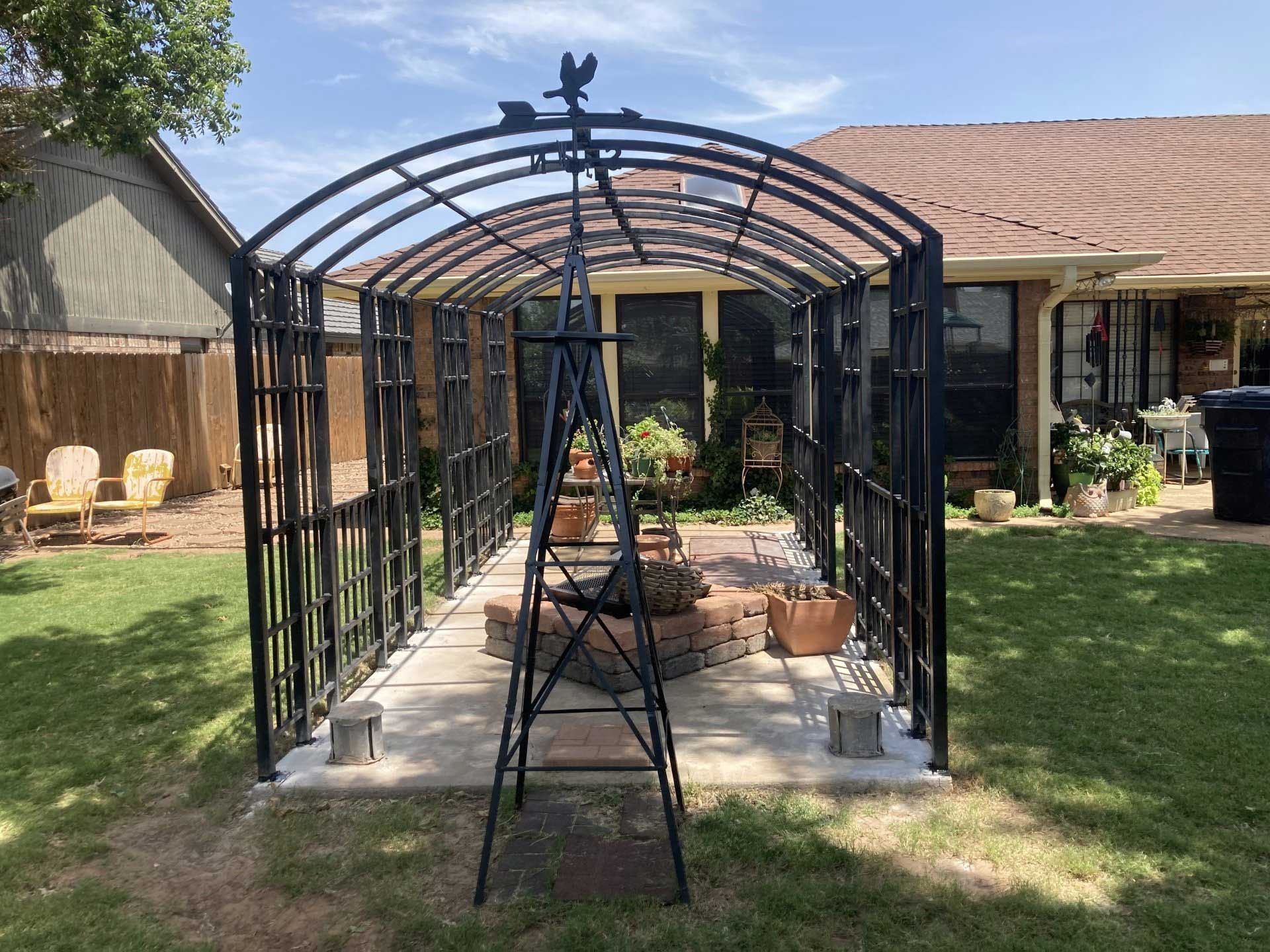 Black metal archway in a backyard, over a concrete path, with a decorative weather vane, and a house in the background.