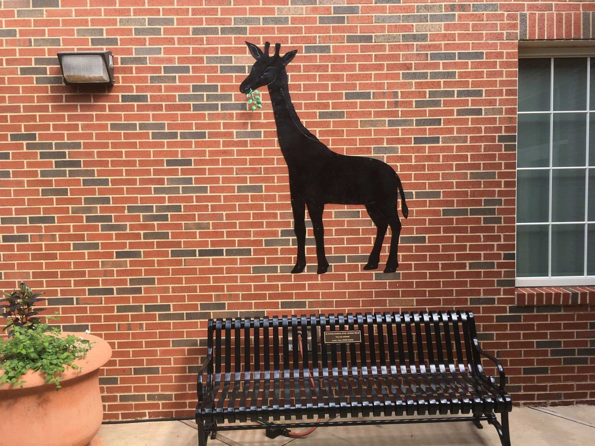 Black metal giraffe sculpture on a brick wall, above a black bench. Plant in a pot to the left.