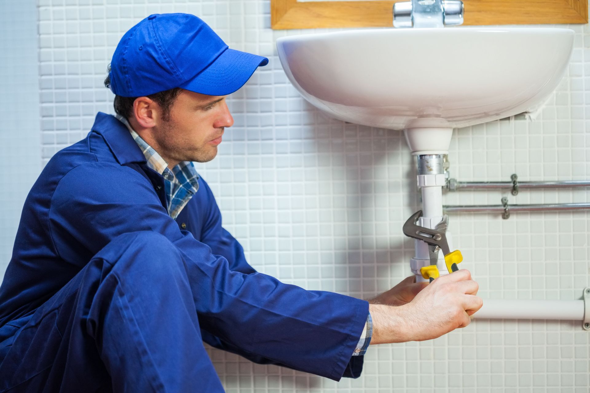 Plumber in blue uniform fixing a sink pipe under a bathroom basin with a wrench