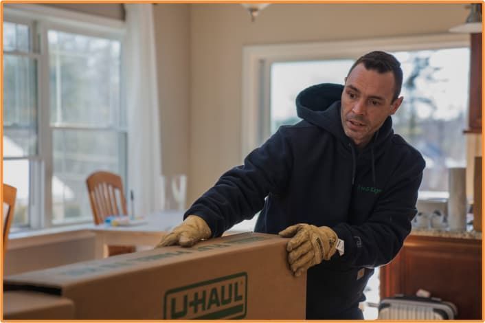 Man in hoodie and gloves carrying a U-Haul box indoors.