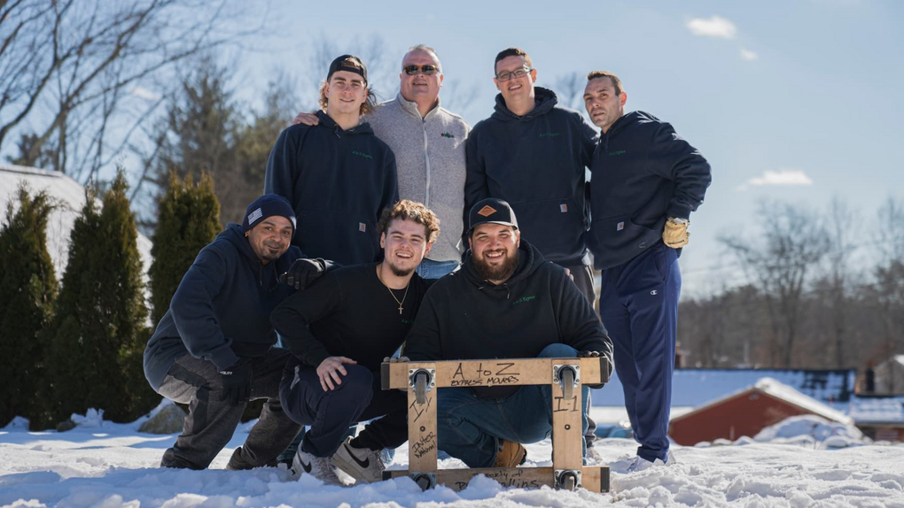 Group of seven men in winter; posing outside. Two men crouch, holding a wooden frame. All wear casual clothes, some smiling.