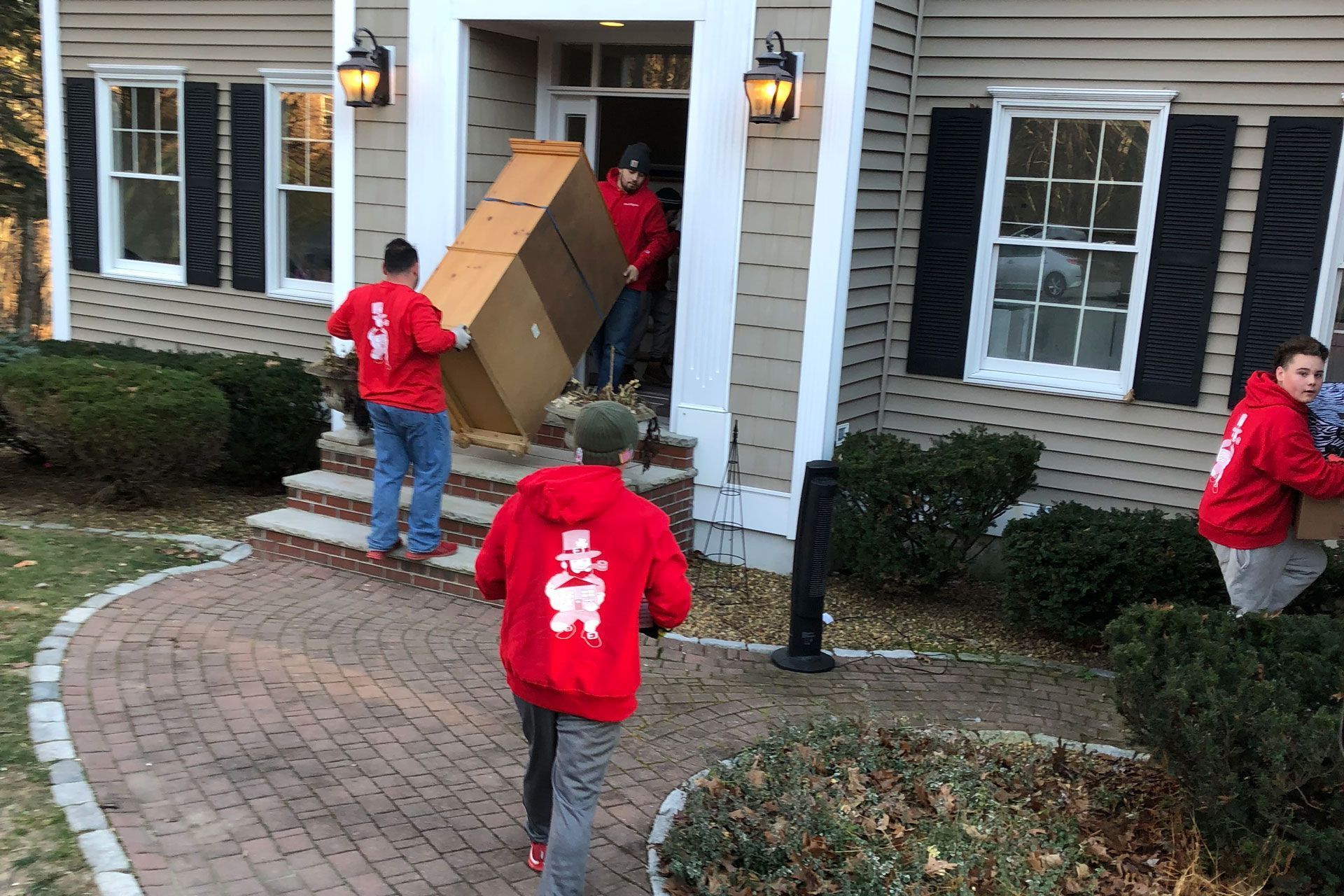 Movers in red hoodies carrying boxes and furniture into a house with a brick walkway.