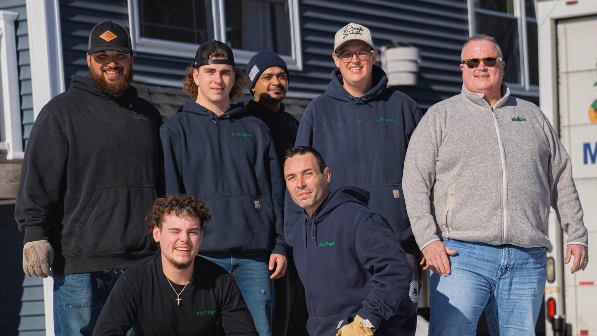 Group of movers smiling in front of a blue building and a truck, wearing work clothes.