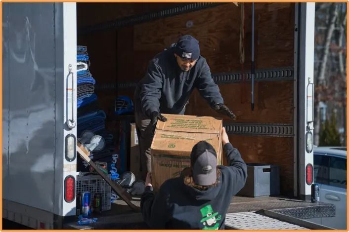 Two people loading a cardboard box into a truck. Outdoors, sunny.