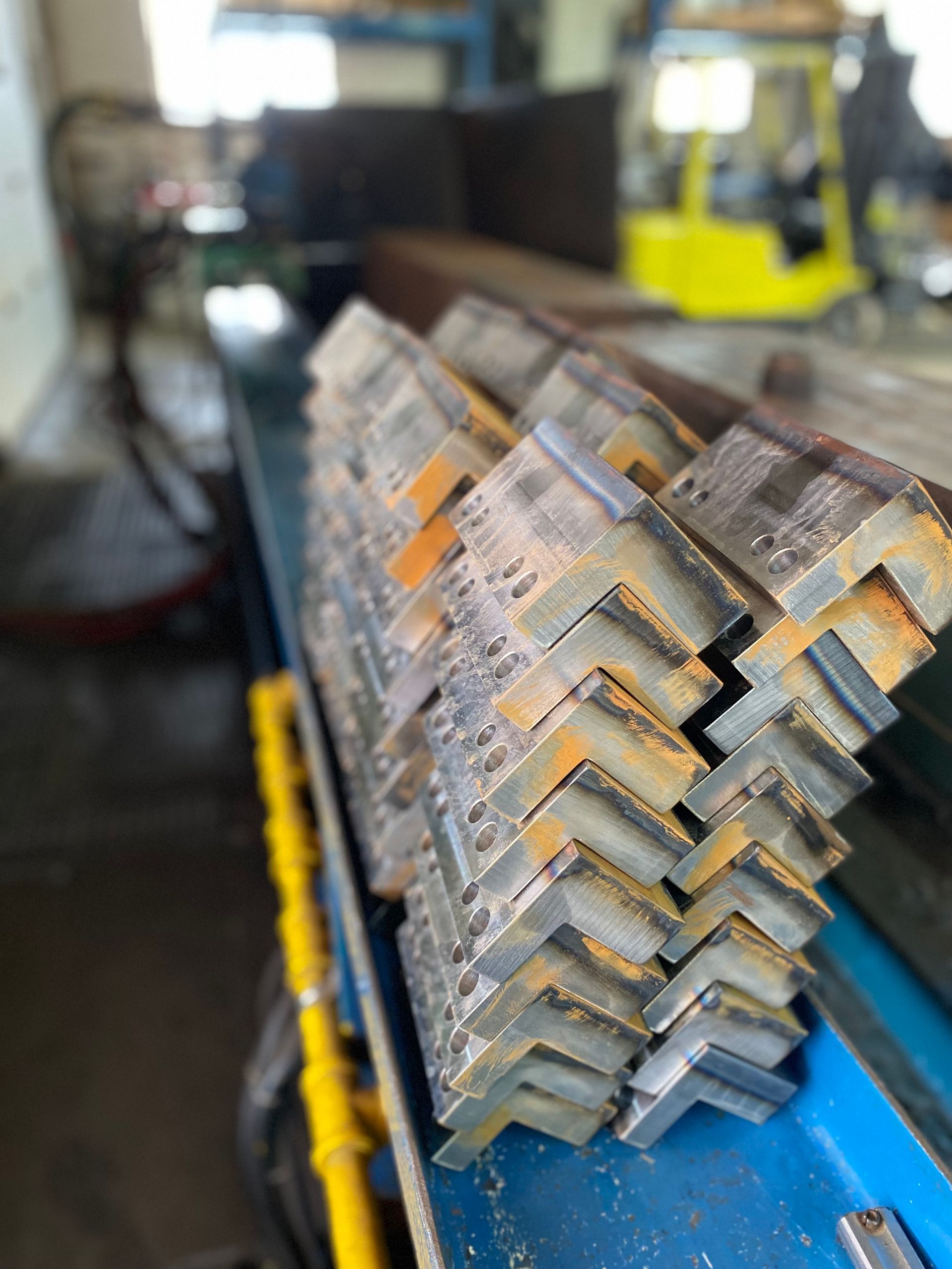 Stack of angle iron pieces in a workshop, rusty edges, close-up shot, some visible machinery in background.