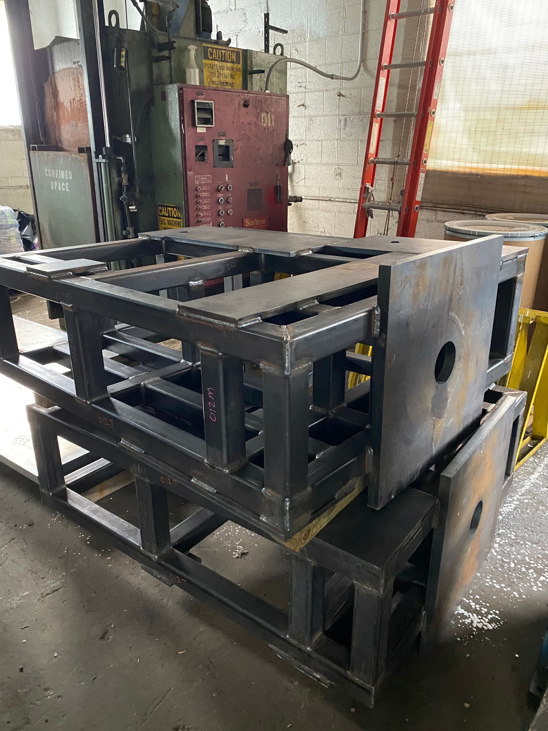 Two large, welded metal structures stacked indoors against a wall with machinery and a ladder.