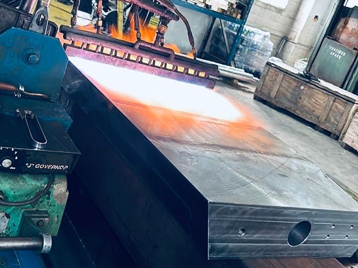 Large, rectangular metal slab being heated with a flame in a factory setting.