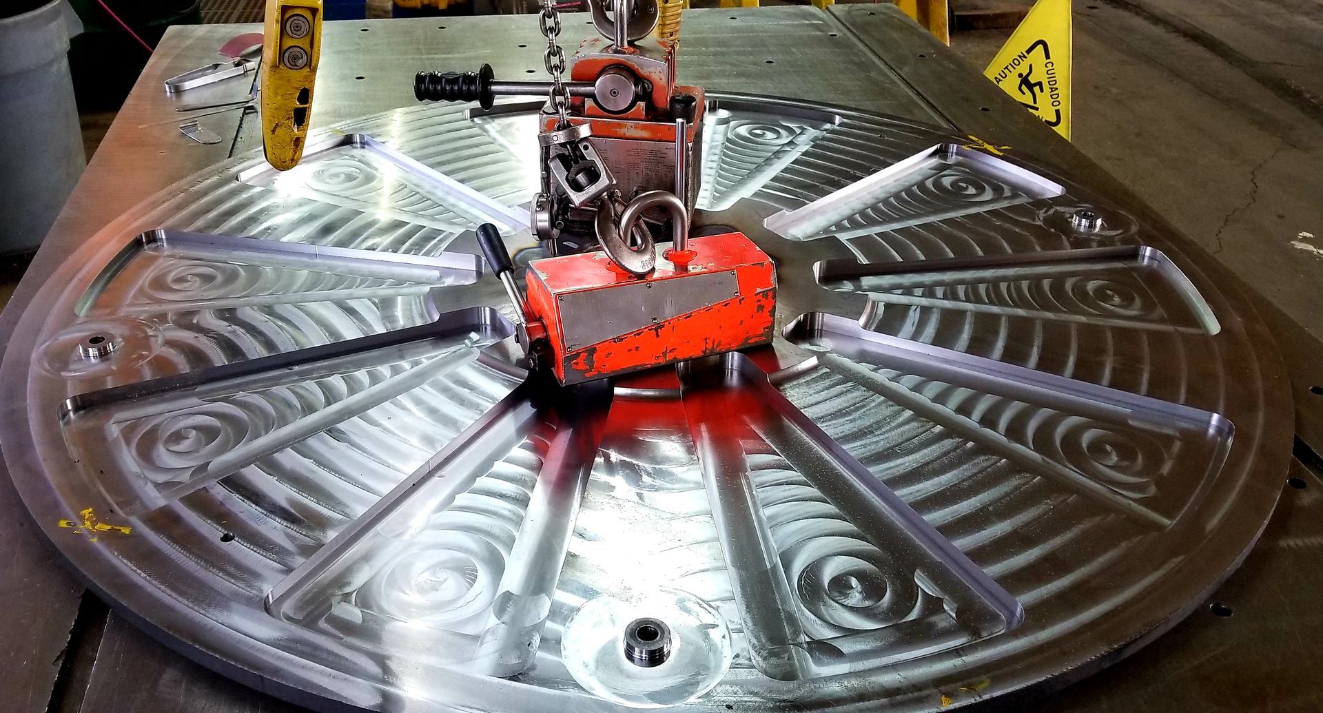 A large, machined metal plate with grooves is being lifted by a crane in a workshop.