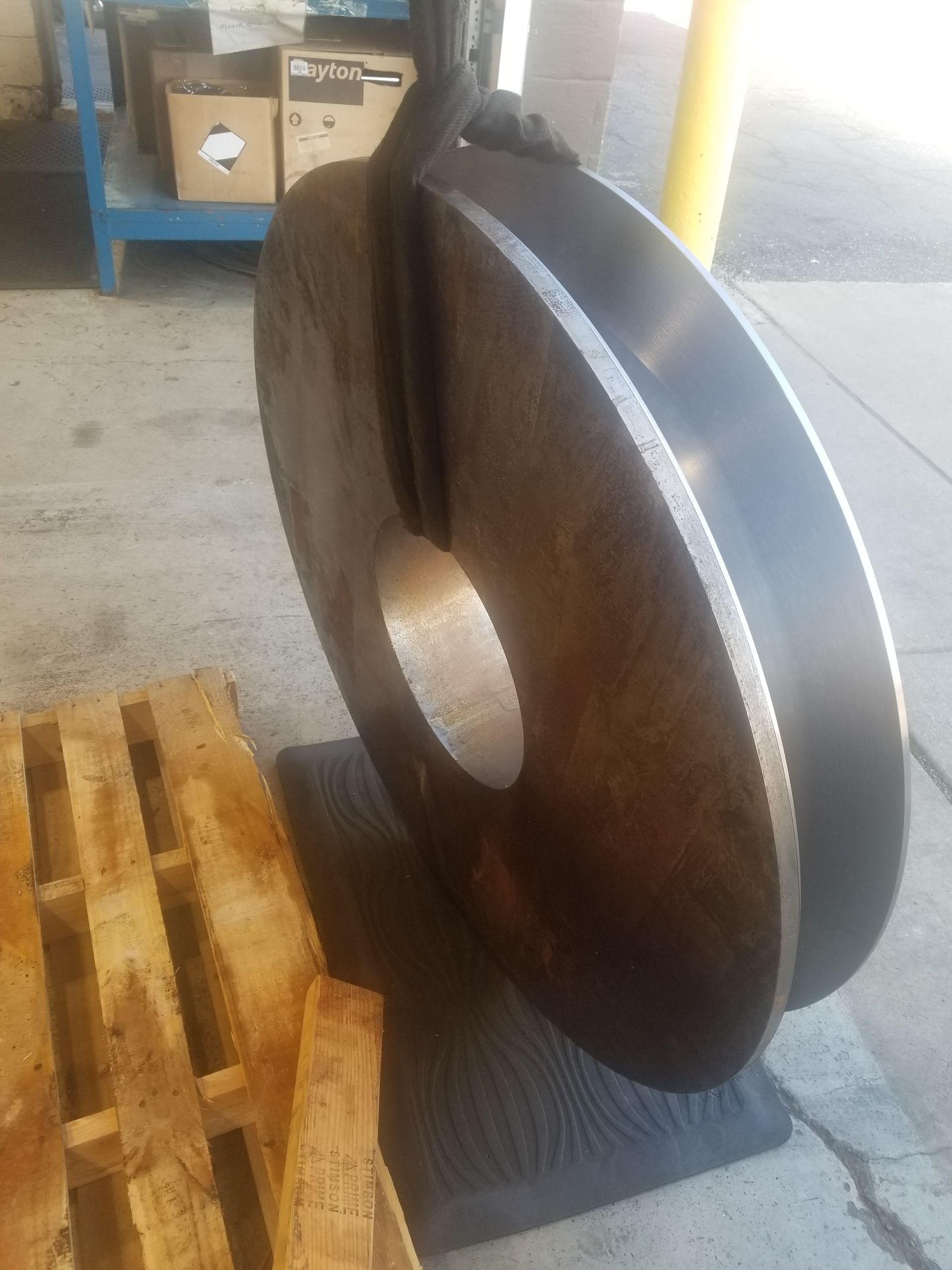 Large metal disk, lifted by a crane hook, resting on a mat near a wooden pallet.
