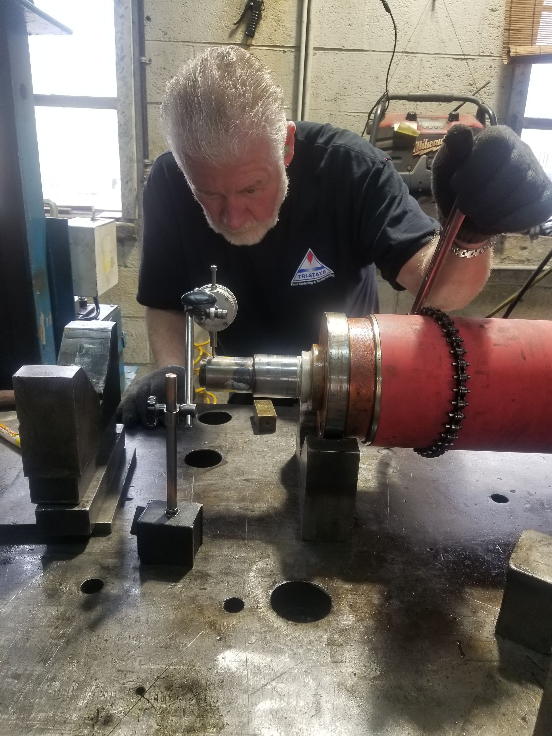 Man using a dial indicator to measure a red cylindrical object on a metal table in a workshop.