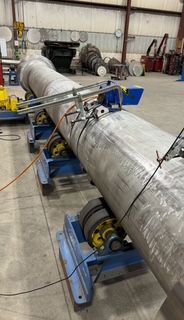 A large metal pipe is being worked on by a machine in a workshop. It sits on blue rollers.