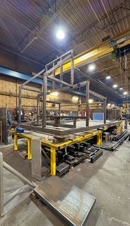 Steel frame structure under construction in a workshop; yellow and blue machinery visible.