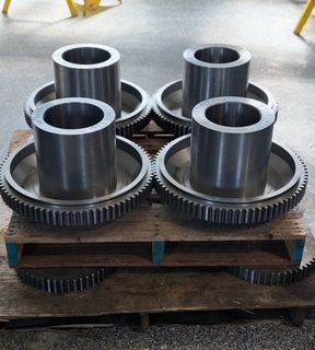 Four large metal gears with cylinders atop, arranged on a wooden pallet.