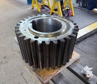 Large industrial gear on a wooden pallet in a workshop. Gray metal, cylindrical with teeth.