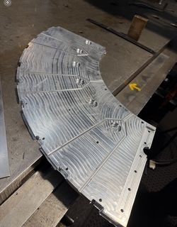 Arc-shaped metal plate with milled grooves and holes, on a metal surface in a workshop.