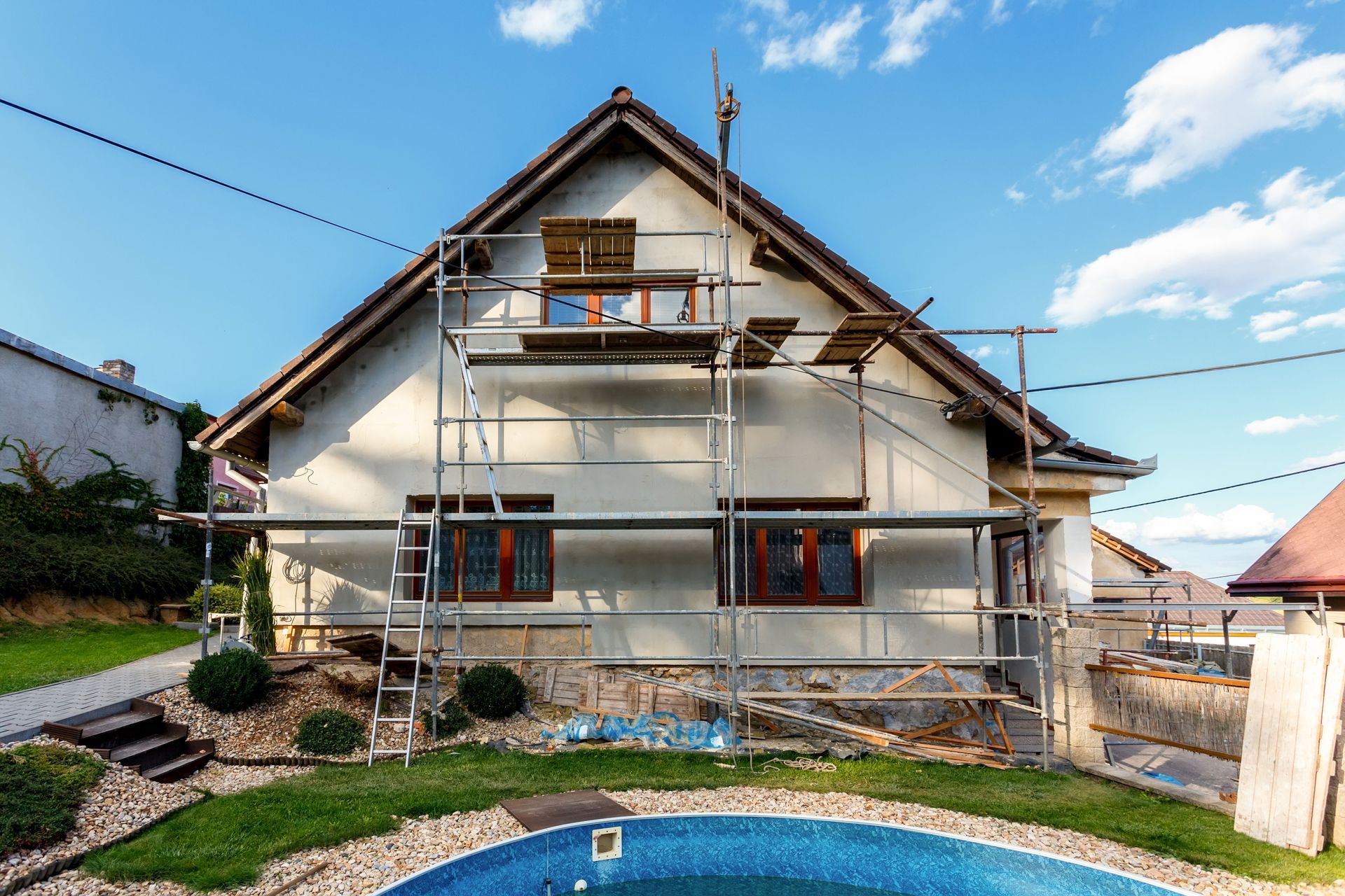 House under renovation with scaffolding, surrounded by grass, pool, and a blue sky.