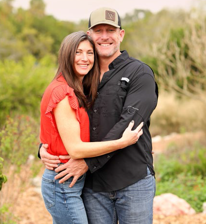 Couple embracing, smiling outdoors. Woman in red top, blue jeans. Man in black shirt, cap.