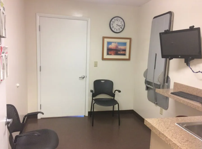 Medical exam room with a chair, clock, and computer screen on the wall.
