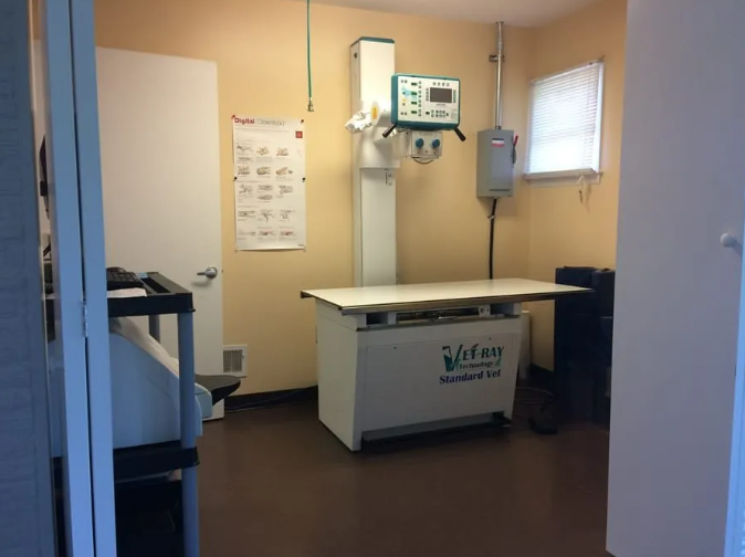 Veterinary clinic x-ray room; tan walls, brown floor, examination table, x-ray machine, white door, and poster.
