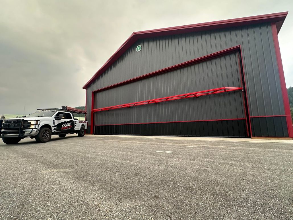 A worker on a lift repairs a large hangar door while two pickup trucks are parked outside.