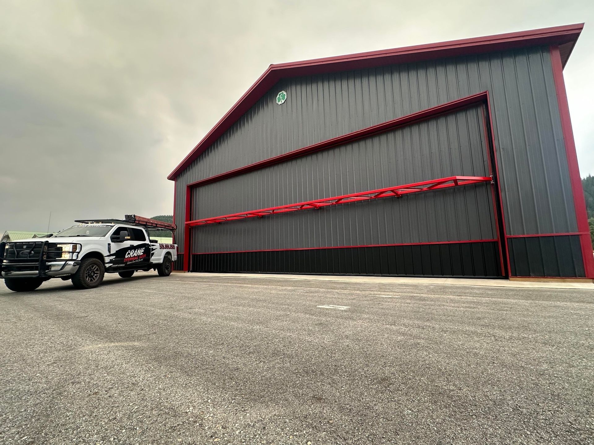 A worker on a lift repairs a large hangar door while two pickup trucks are parked outside.