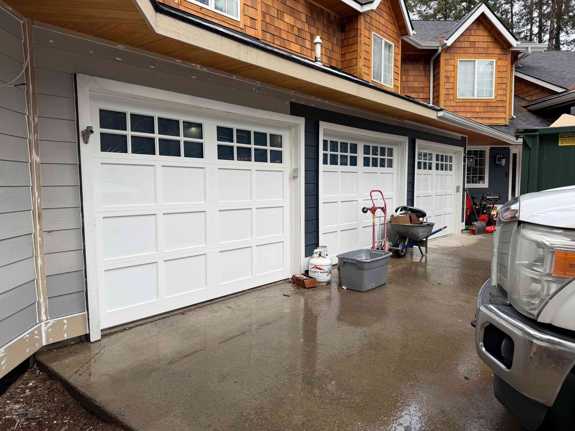 A exterior view of a house with a three-car garage, white doors, gray siding, and cedar shake accents on a rainy day.