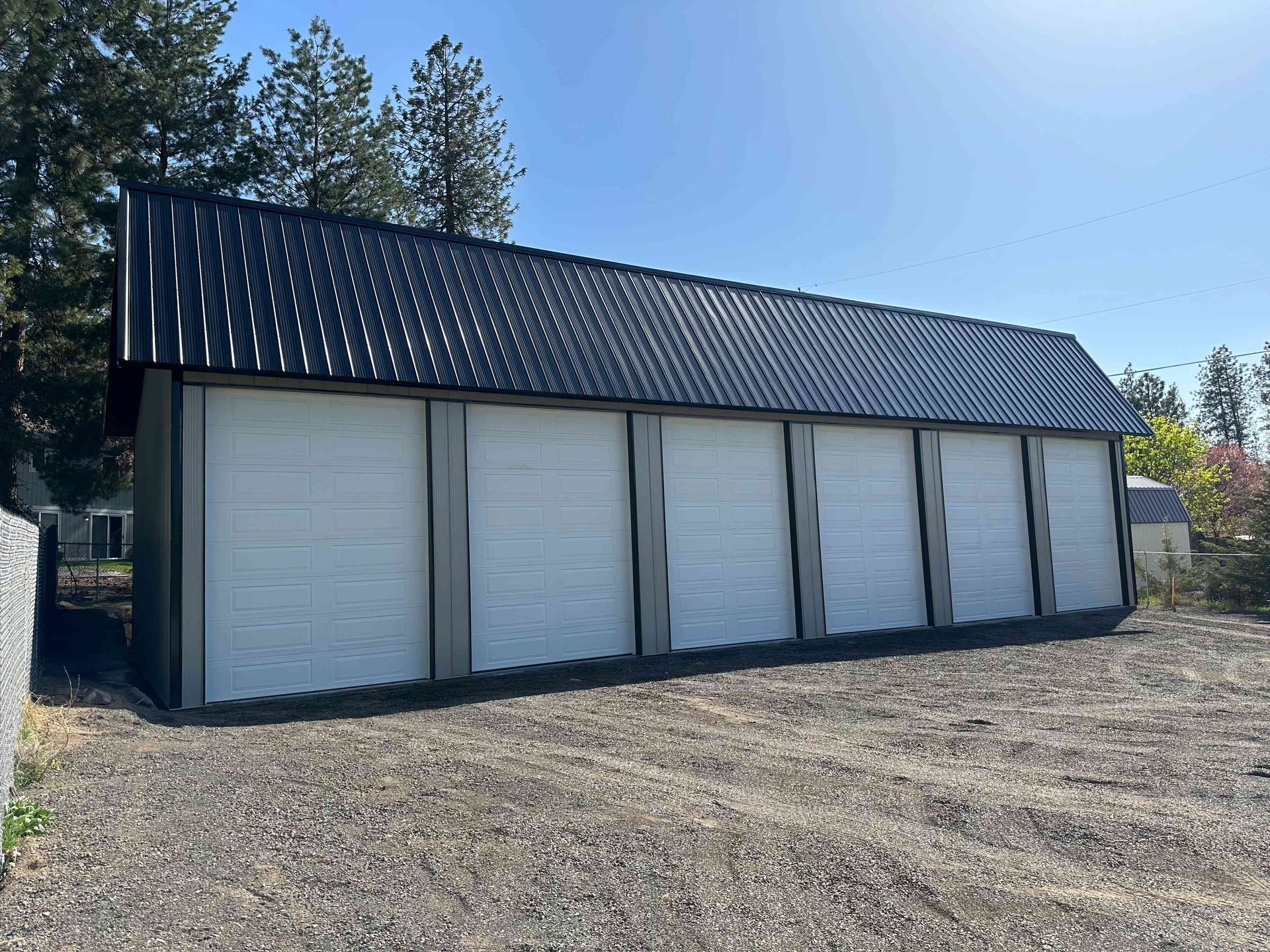 A long, rectangular storage building with a black metal roof and six white roll-up garage doors on a gravel lot.
