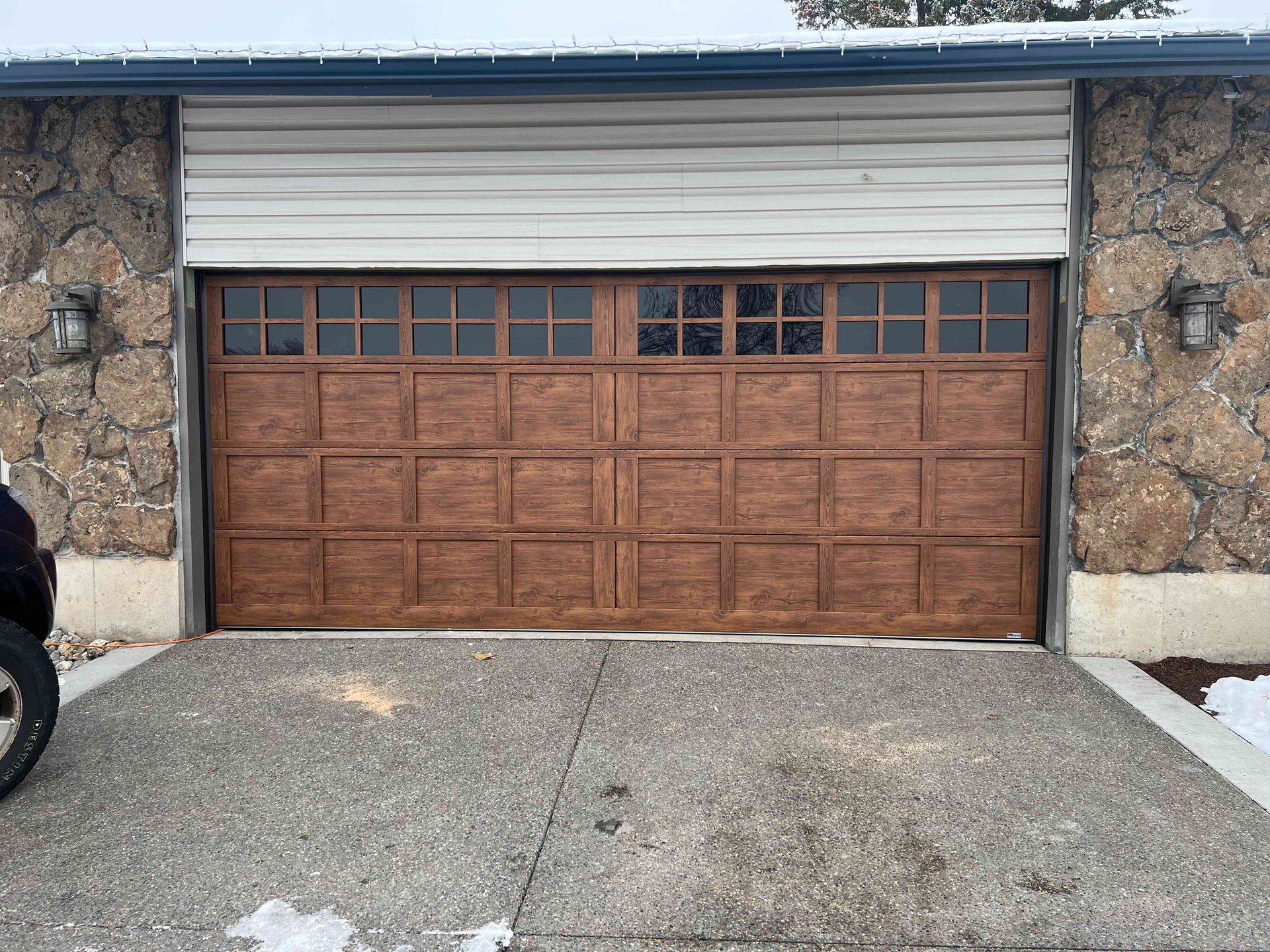 A brown garage door with rectangular panels and a row of windows set into a stone wall house exterior.