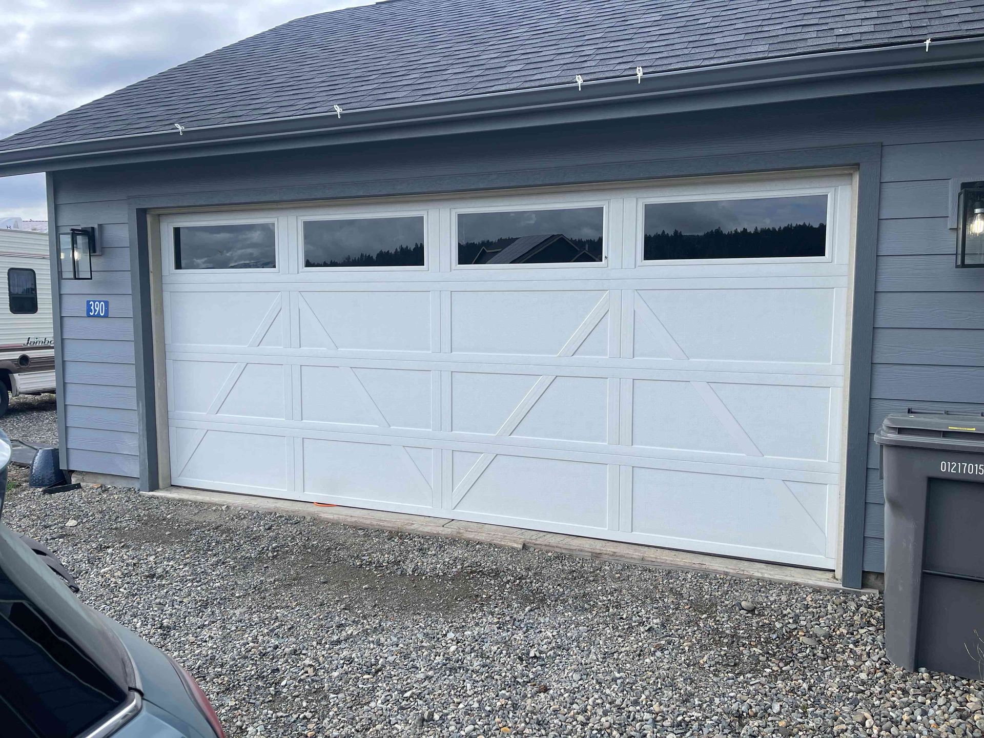 A white, paneled garage door with top row windows installed on a grey building exterior with a gravel driveway.