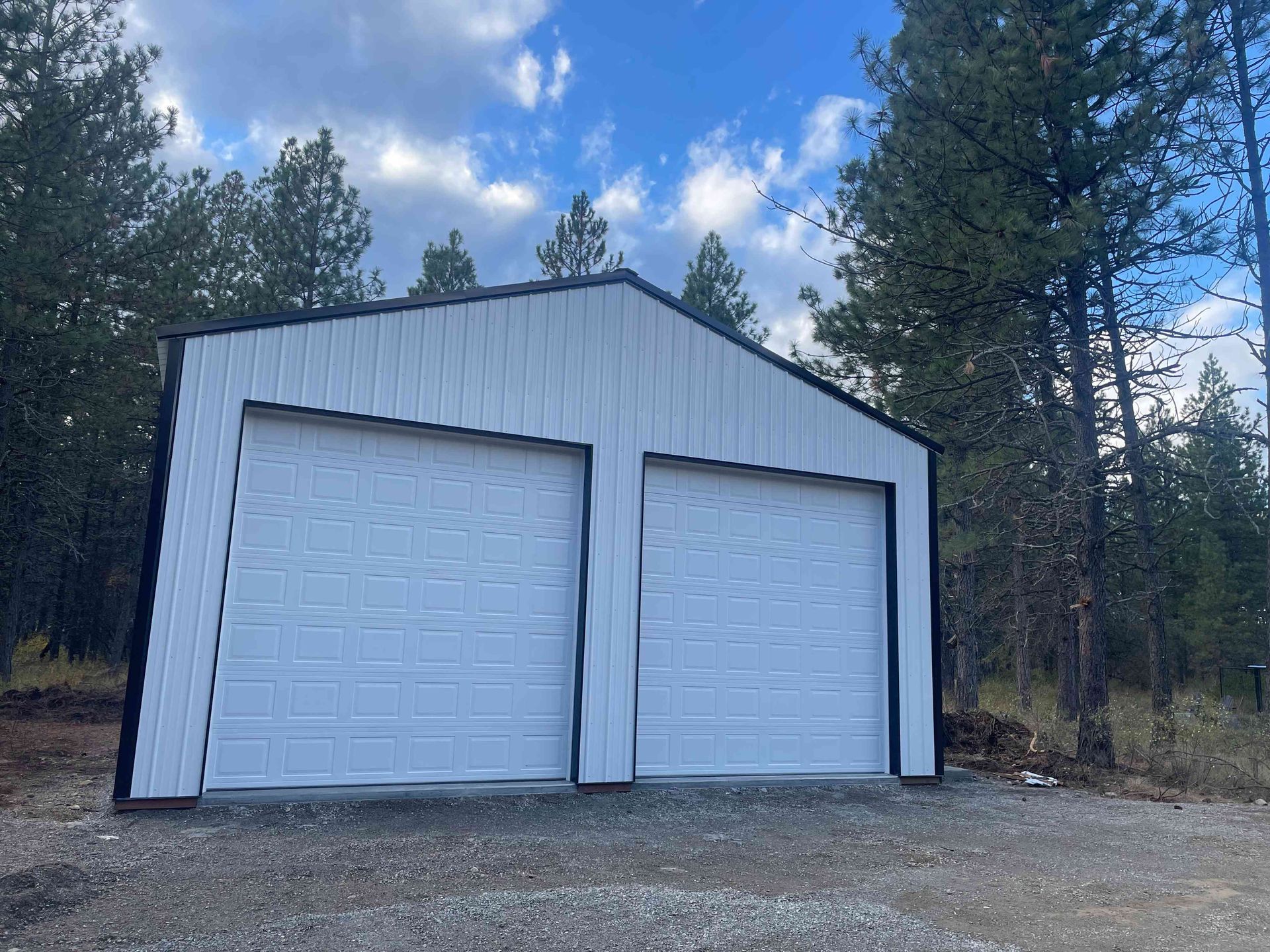 A modern white, two-car metal garage building with black trim, situated in a wooded area under a blue sky.