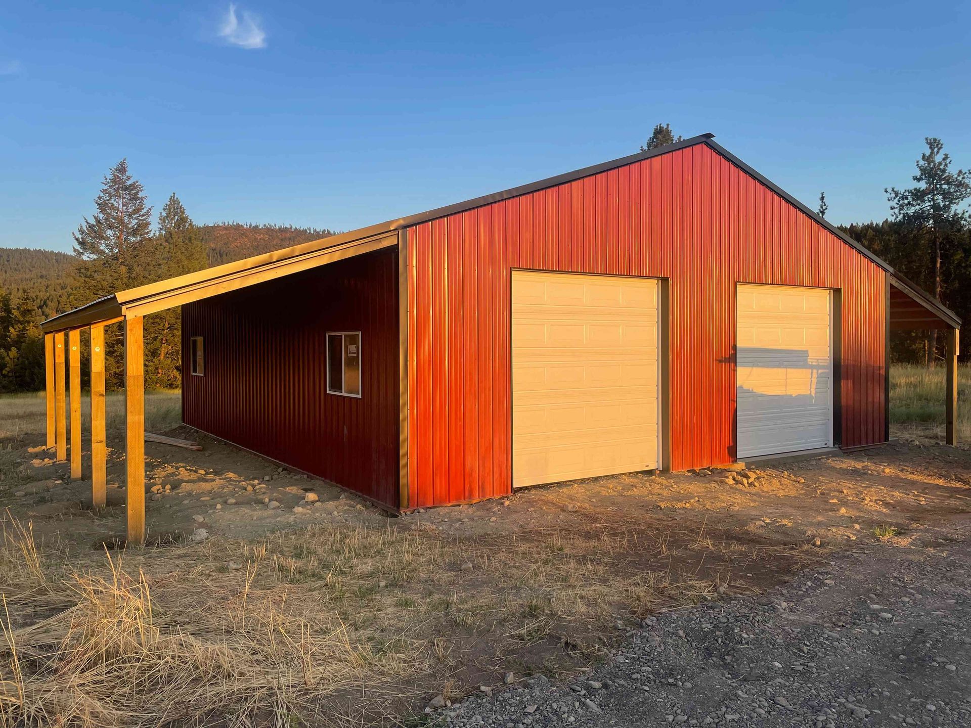A red metal barn with two garage doors and an open lean-to porch sits in a rural, grassy area under a blue sky.