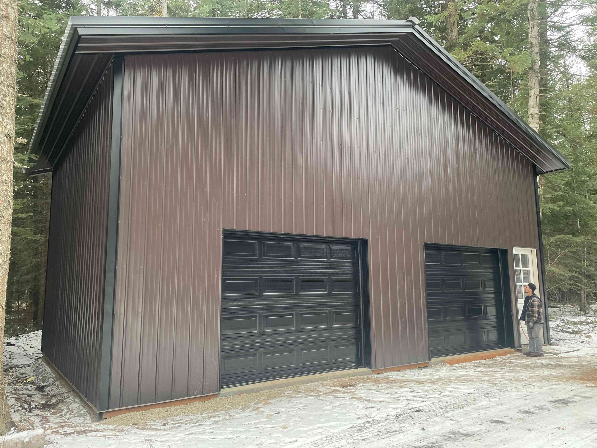 A brown metal two-car garage with black roll-up doors, set in a snowy, wooded area with a person standing by the door.