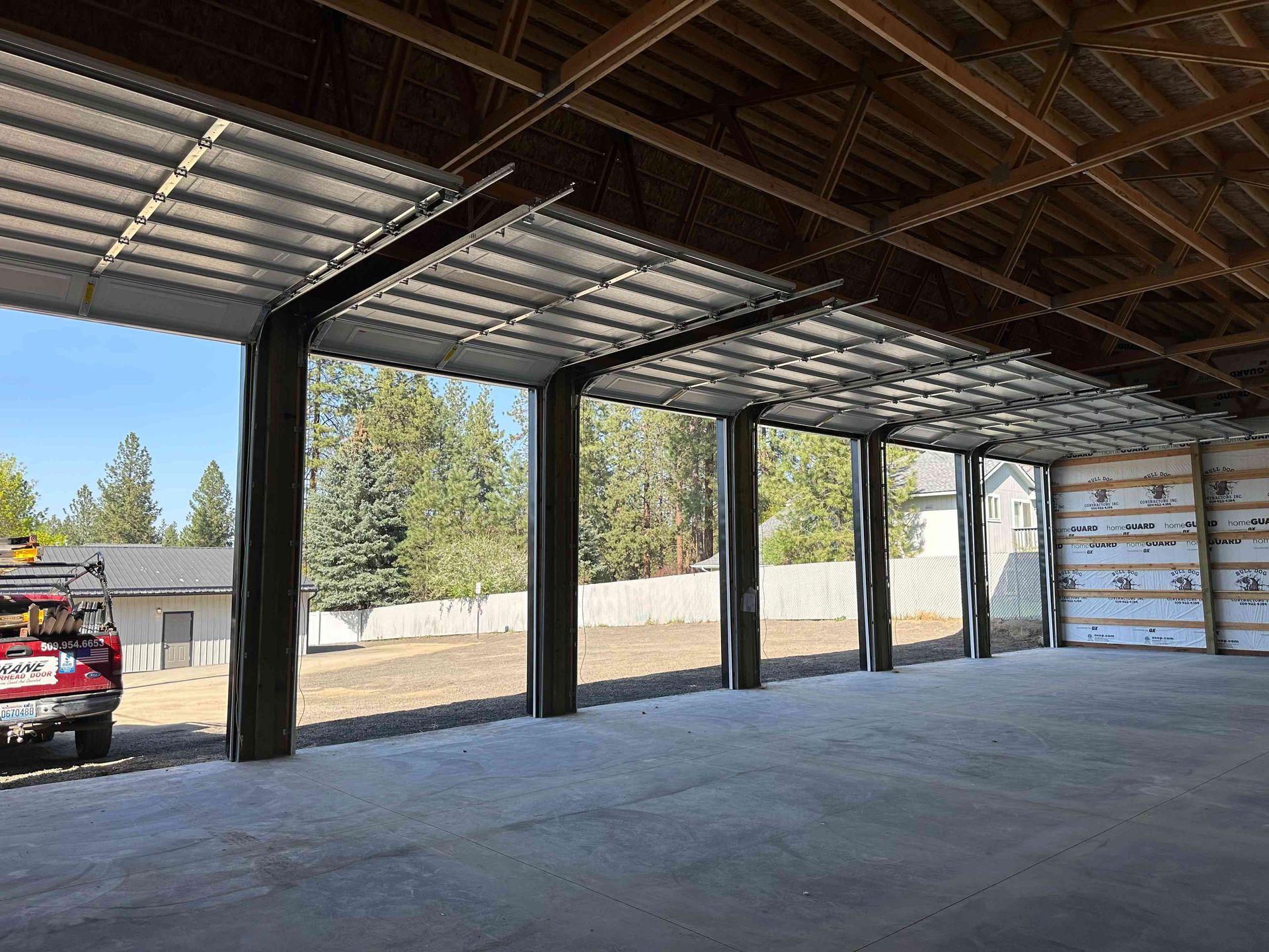 A spacious, unfinished workshop interior featuring open garage bays revealing a sunny outdoor lot with trees and a house.