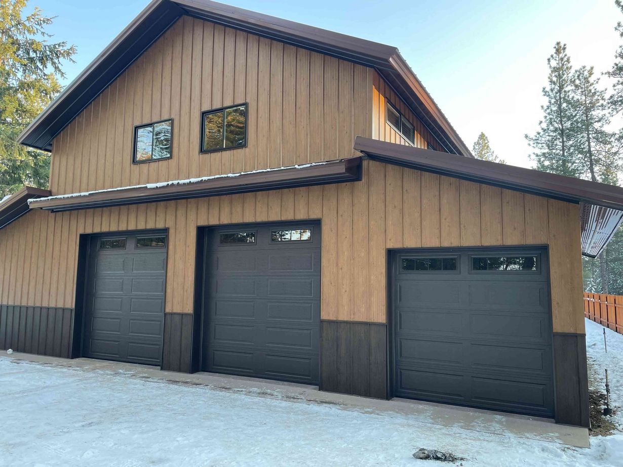 A two-story garage with board-and-batten tan siding, dark trim, three garage doors, and snow on the ground.