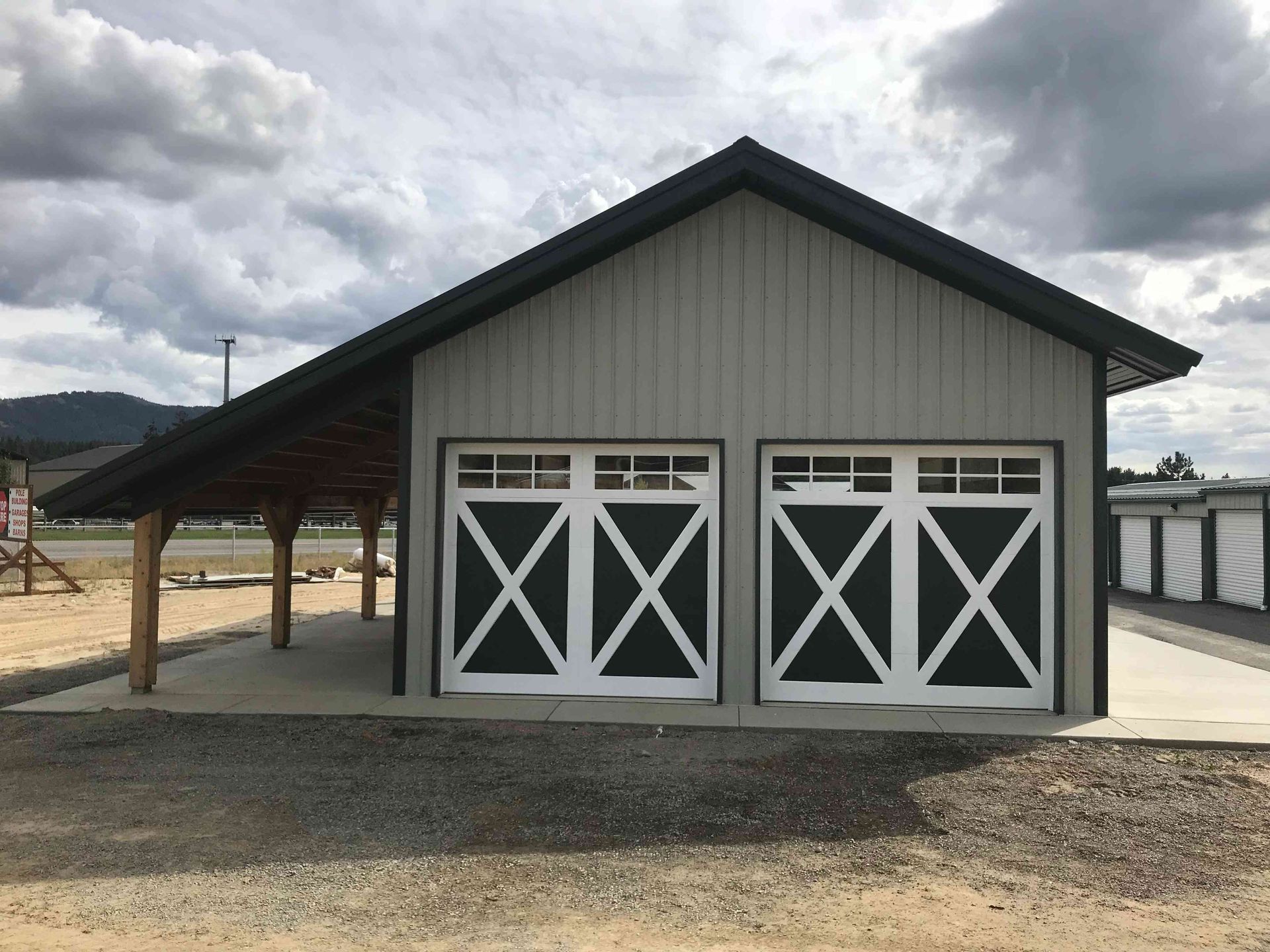 A beige metal barn with two dark-trimmed garage doors and an attached open-air wood porch under a cloudy sky.