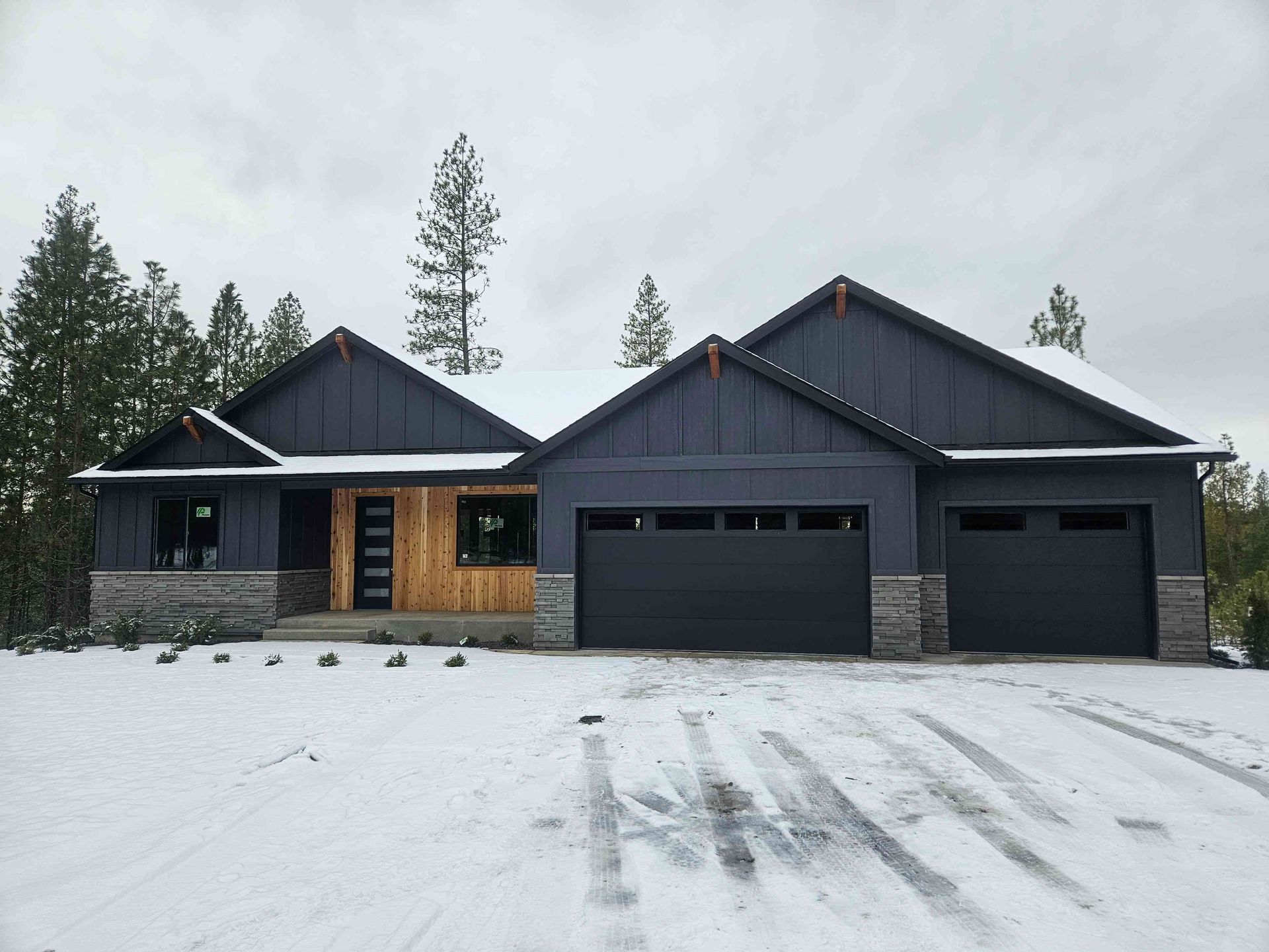 A modern, dark-gray house with vertical siding and stone accents, featuring a wood-paneled entryway, covered in light snow.
