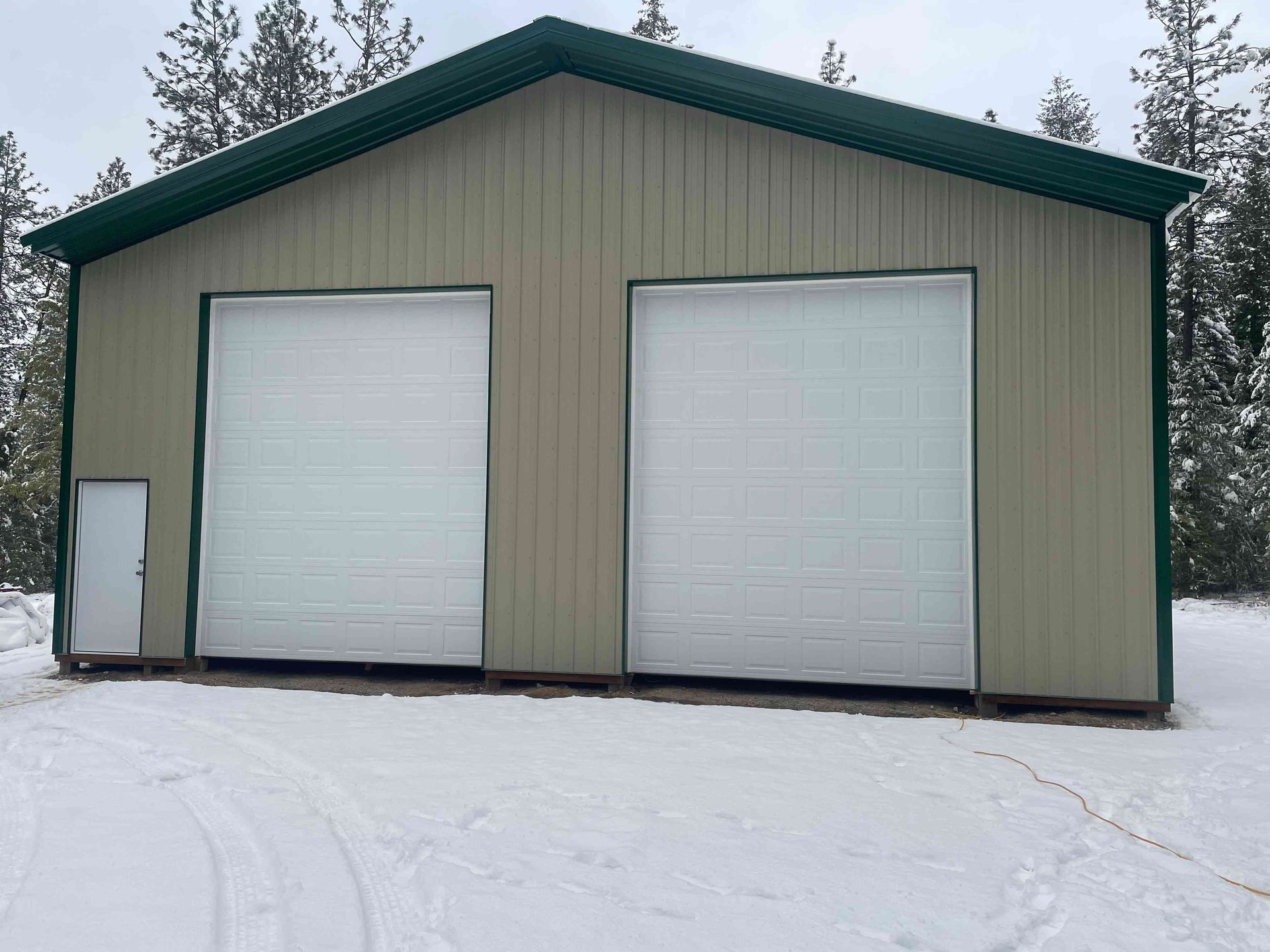 Tan-colored pole barn with a green roof, two white garage doors, and a small side door, set in a snowy, wooded area.