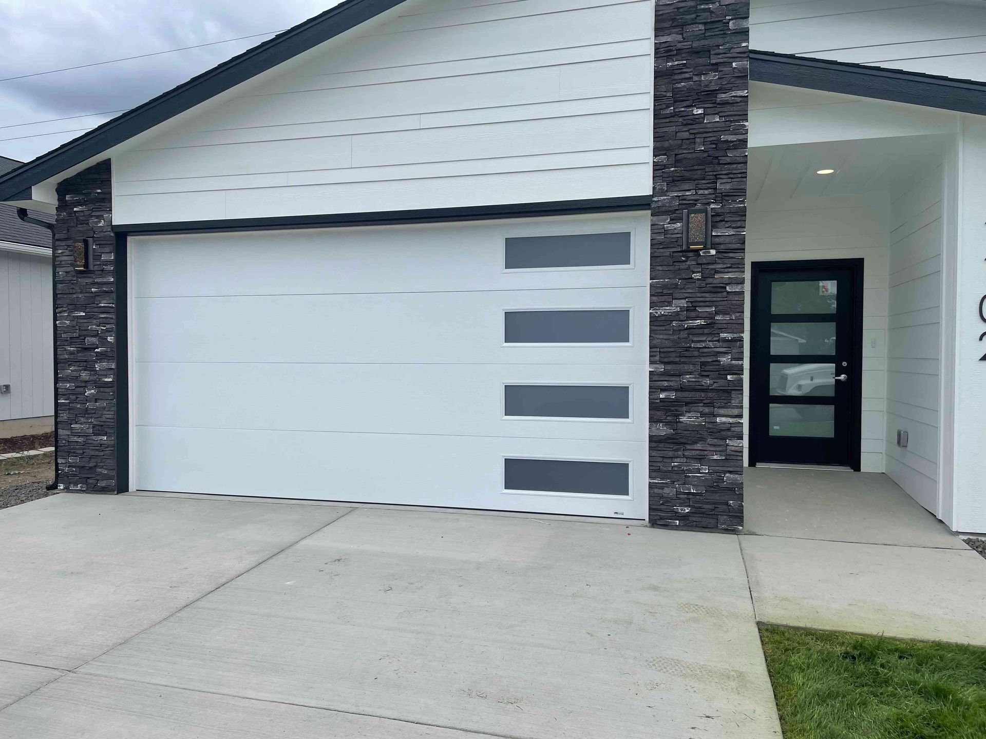 White garage with black stacked stone accents and a modern black-framed glass front door on a concrete driveway.