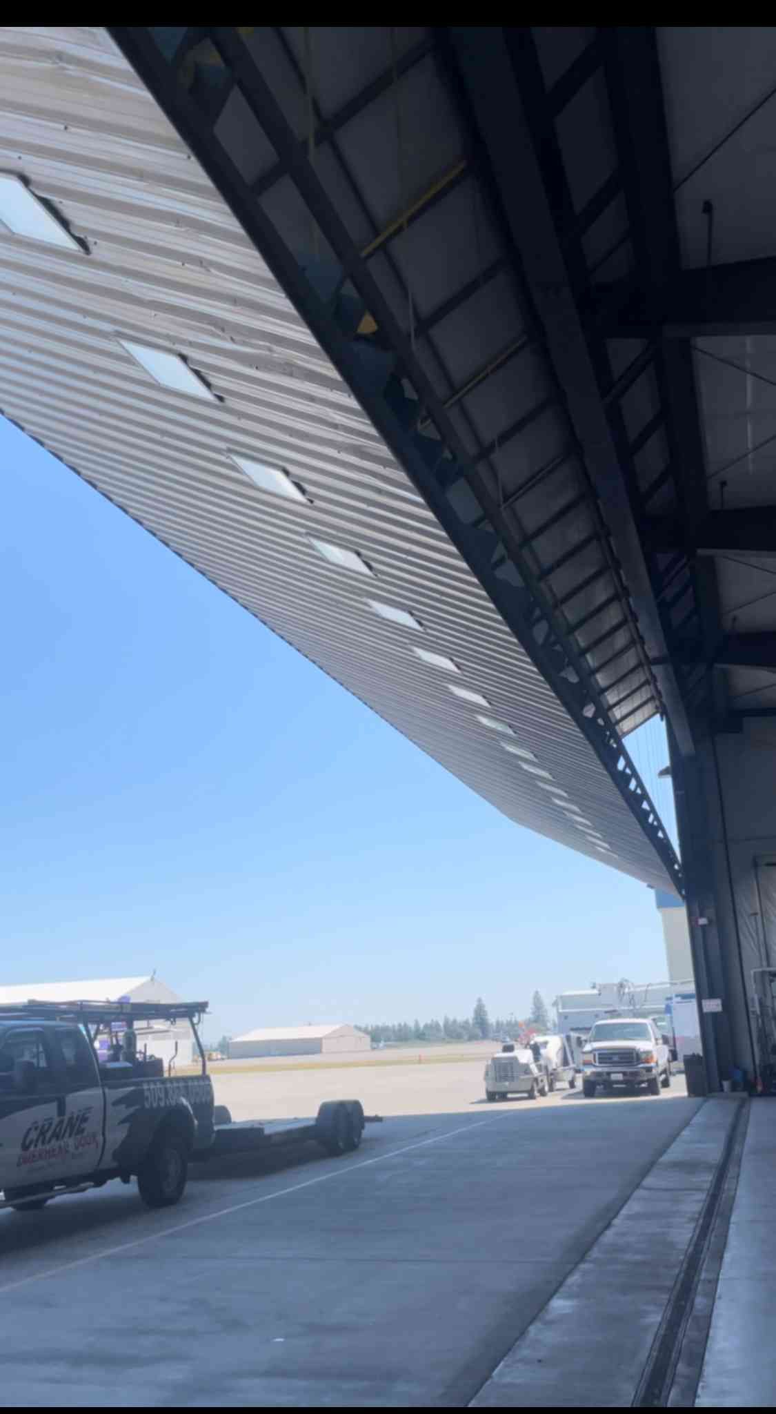 Looking out from beneath a large hangar roof at an airfield with parked vehicles under a clear blue sky.