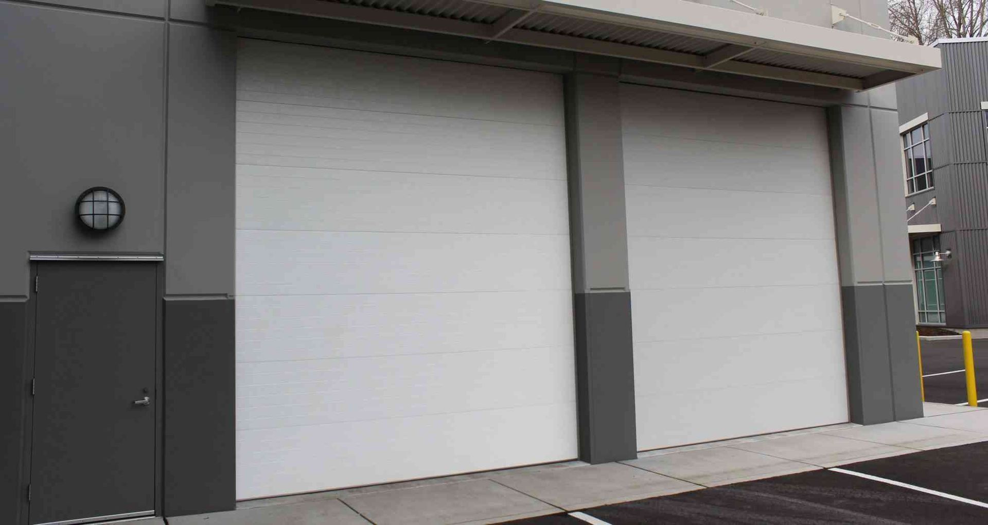 A dark gray commercial building exterior with a single entrance door and two large, white roll-up garage doors.