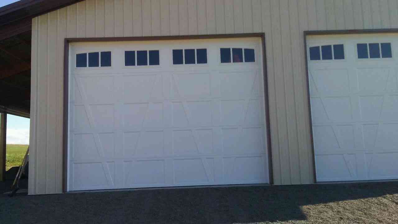 Two white garage doors with rectangular windows on each, set in a light beige building with a gravel driveway.