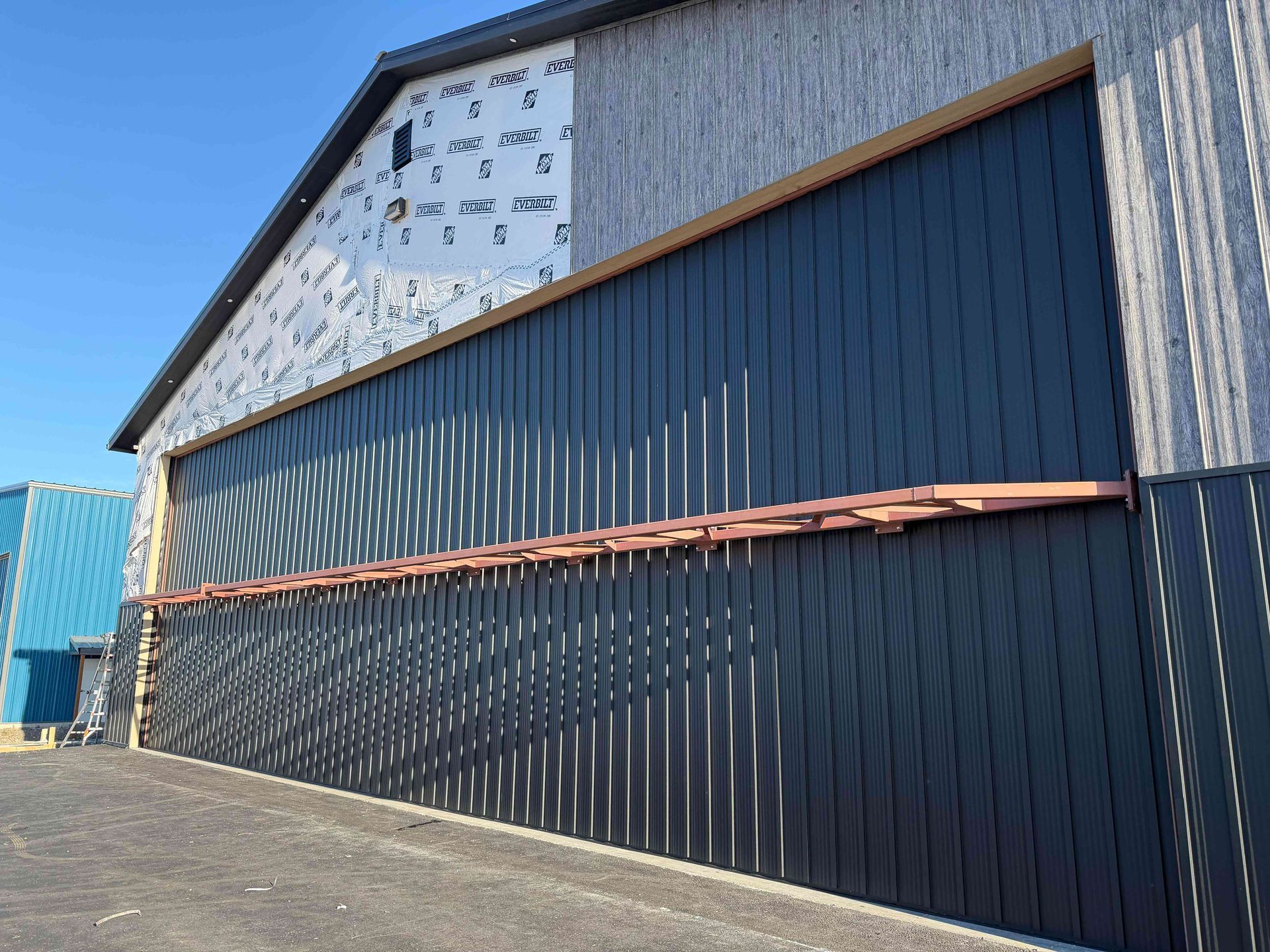 A large, dark metal building facade with vertical siding under a clear blue sky, partially covered in protective wrap.
