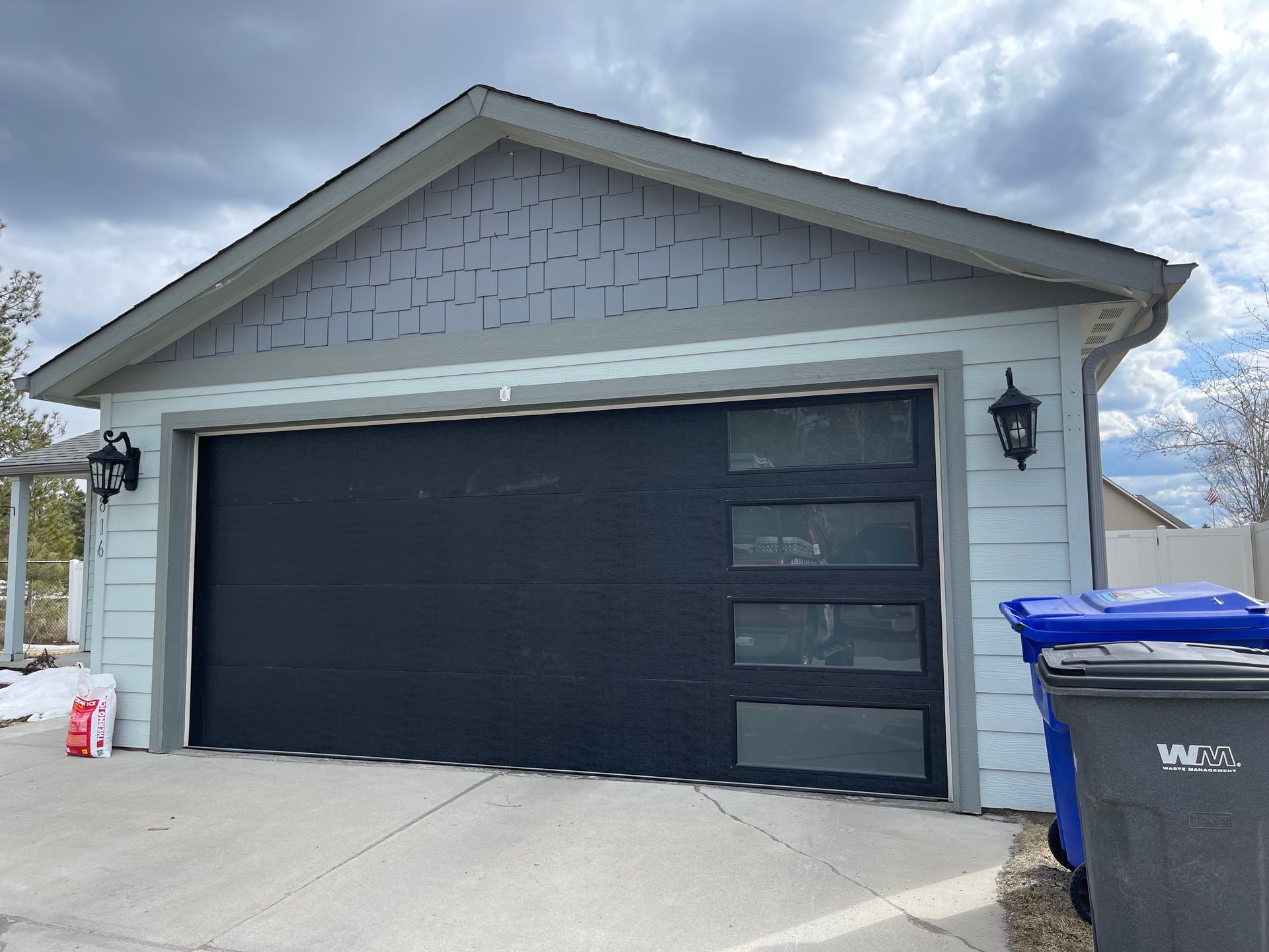 A detached garage with light blue siding, a gray shingle gable, and a black garage door with four vertical glass windows.