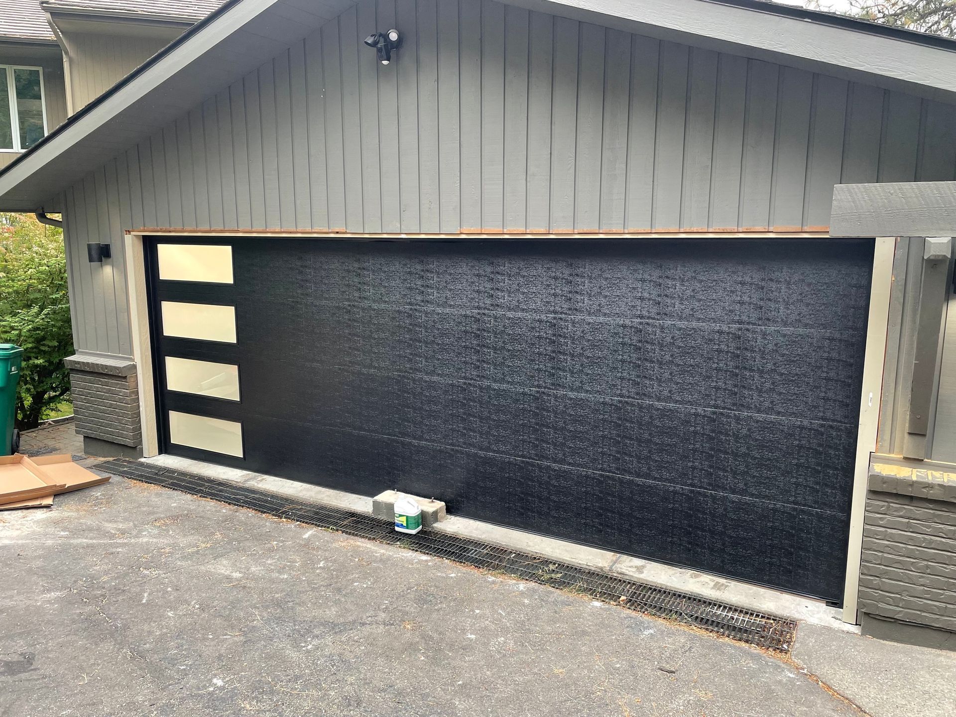 A modern, black-painted garage door with four vertical frosted windows on the left, set in a grey house exterior.