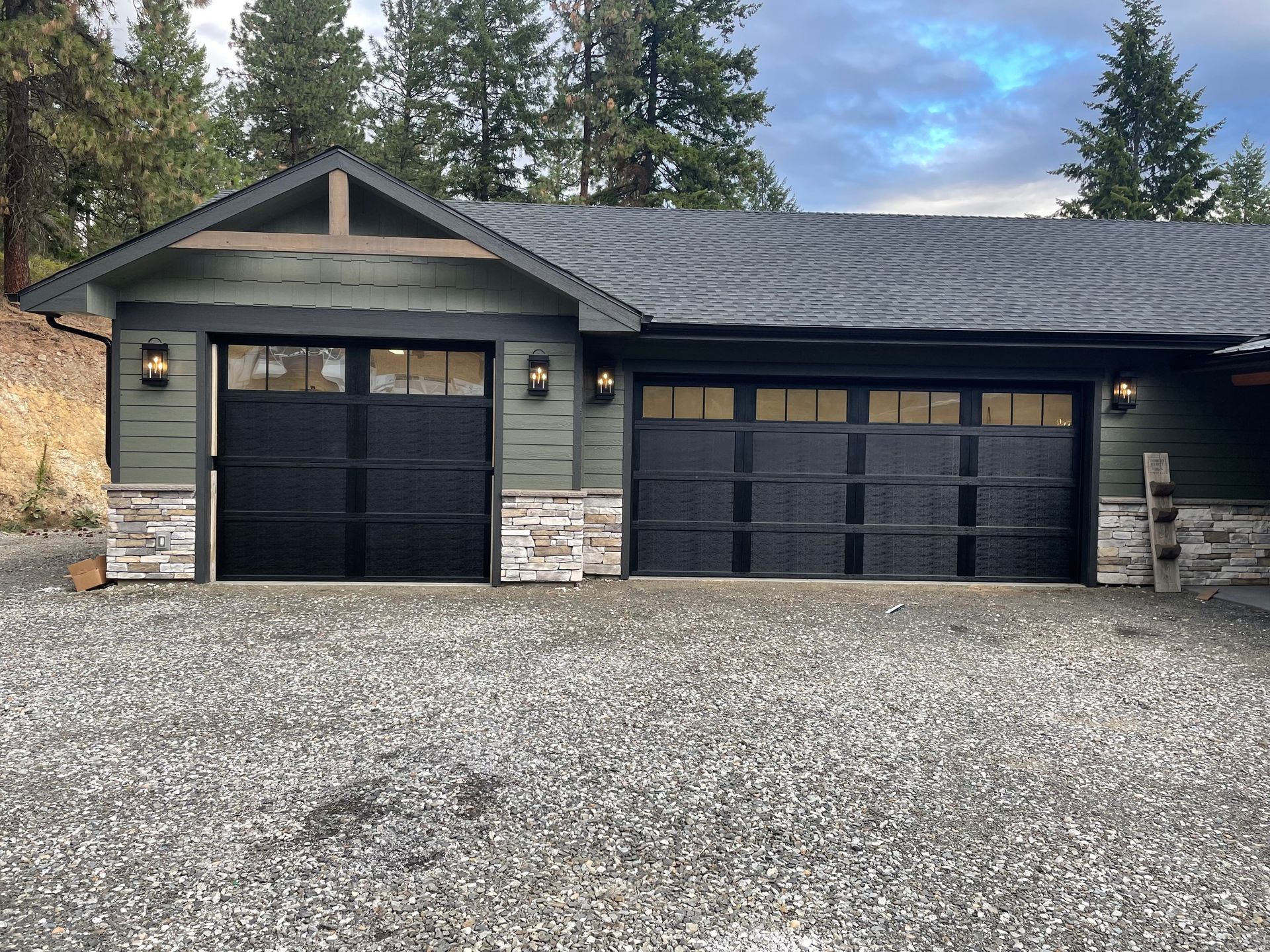 A two-car garage with dark green siding, stone accents, black doors with windows, and a shingled roof in a wooded area.