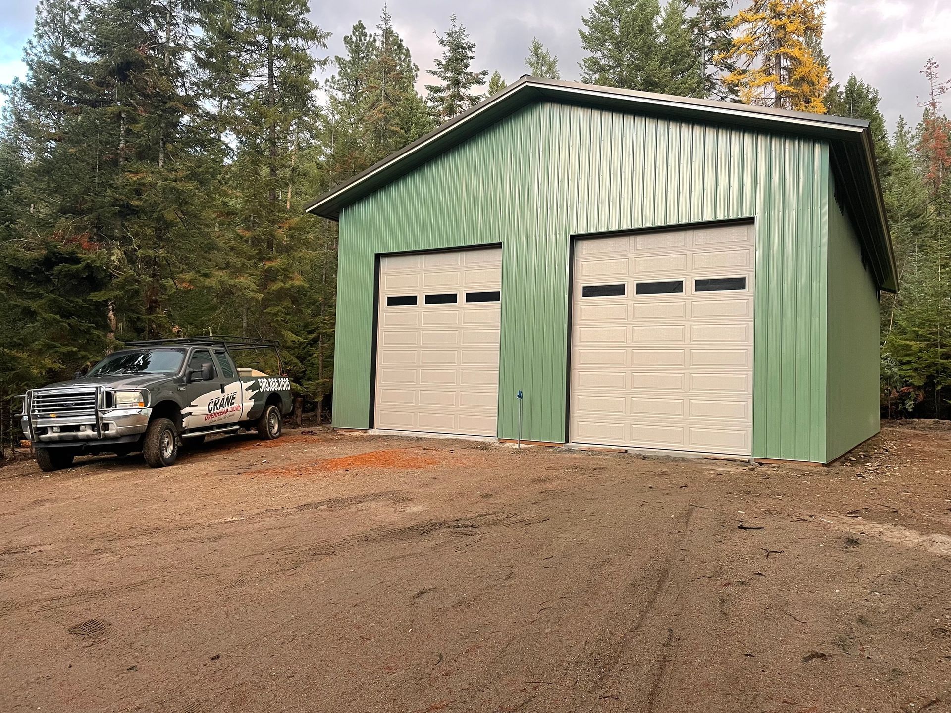 A green metal garage building with two white doors sits in a wooded area, with a pickup truck parked to the left.