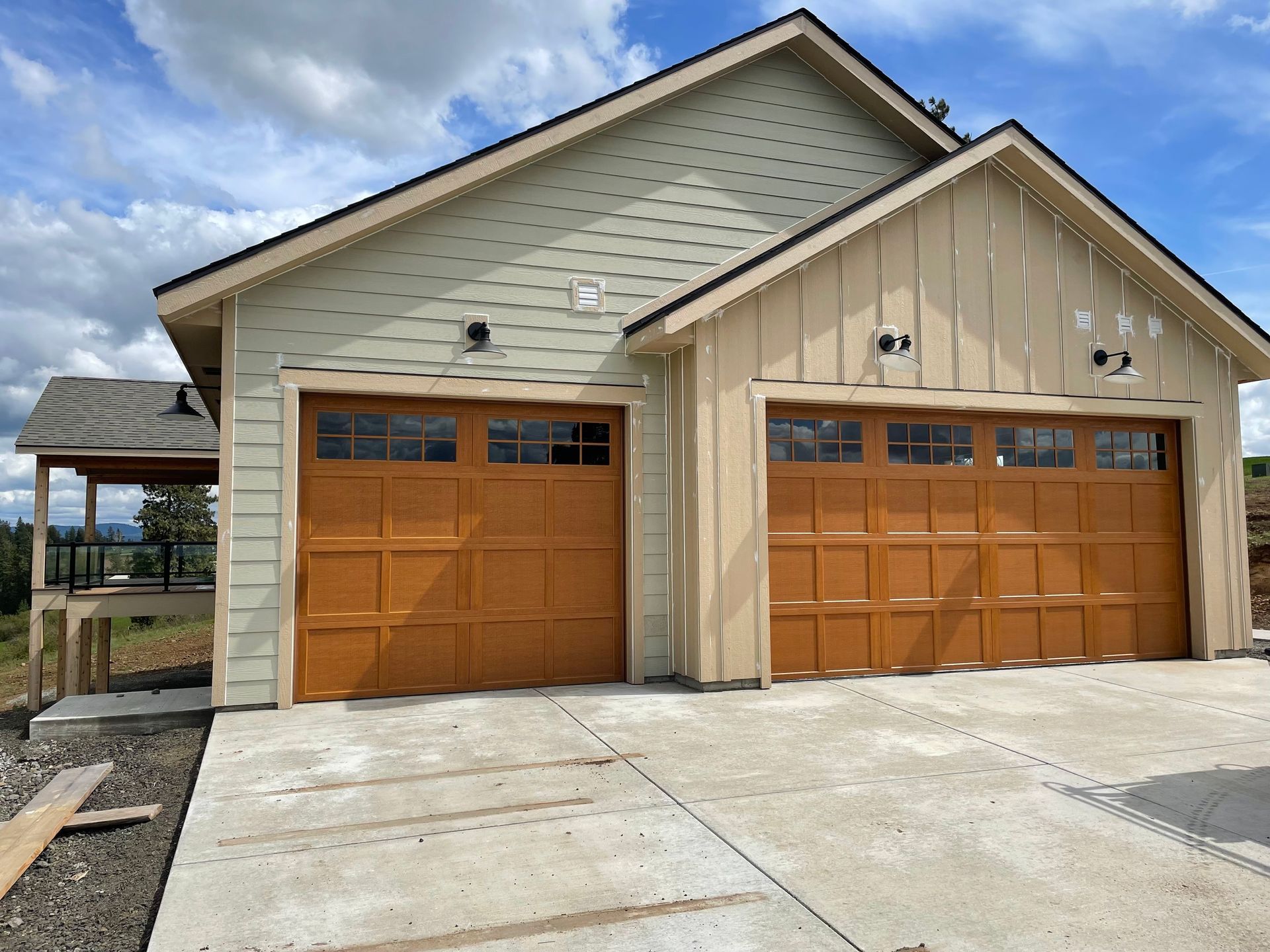 A two-car garage with tan siding and brown doors, featuring a side porch, set against a blue sky with clouds.