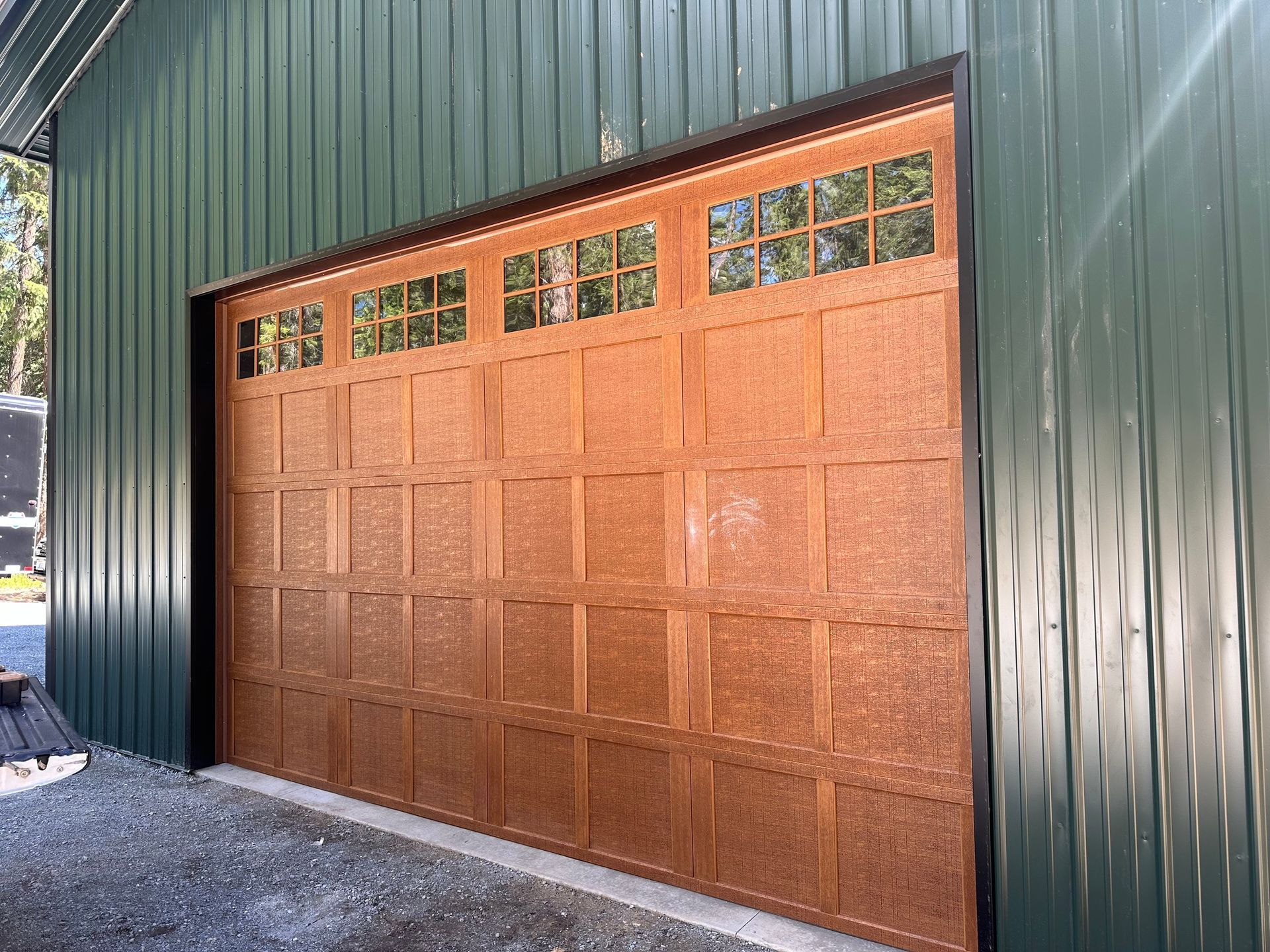 A brown, wood-paneled garage door with top window inserts, installed on a building with vertical dark green metal siding.
