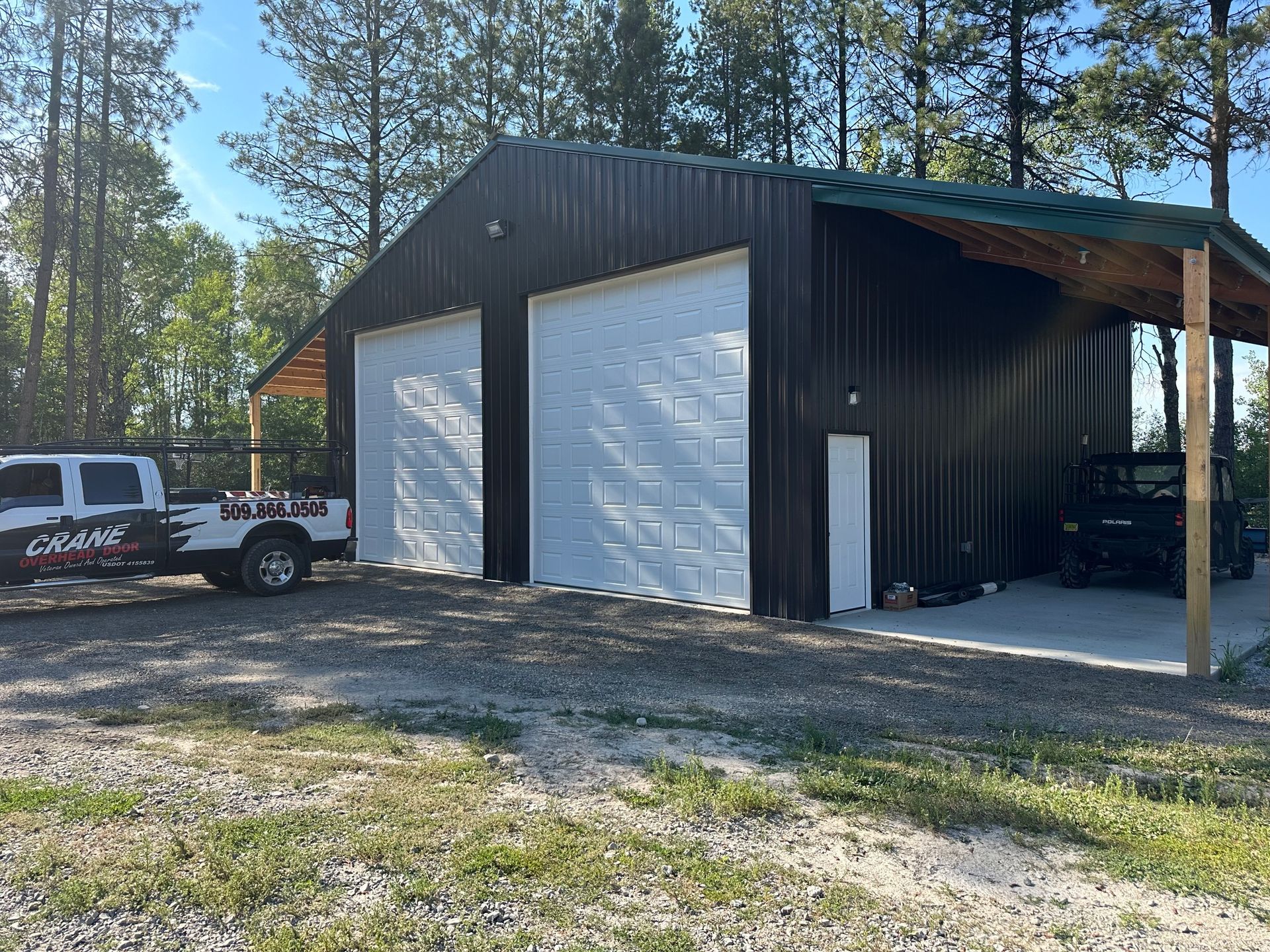 A dark-walled metal workshop with two white garage doors, a side entry, and an open lean-to shelter next to a parked truck.
